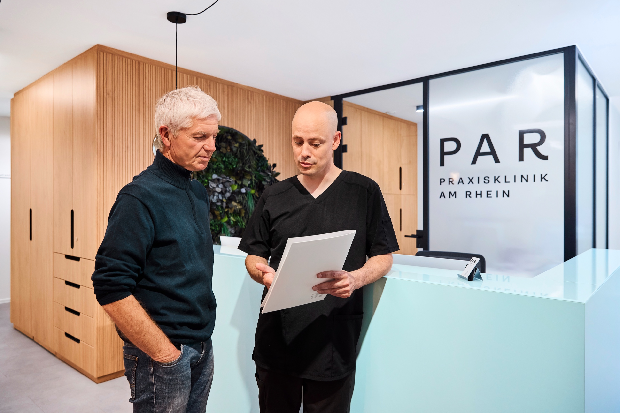 Dr. Romain Doliveux erläutert einem Patienten ein Dokument am Empfang der Praxisklinik am Rhein